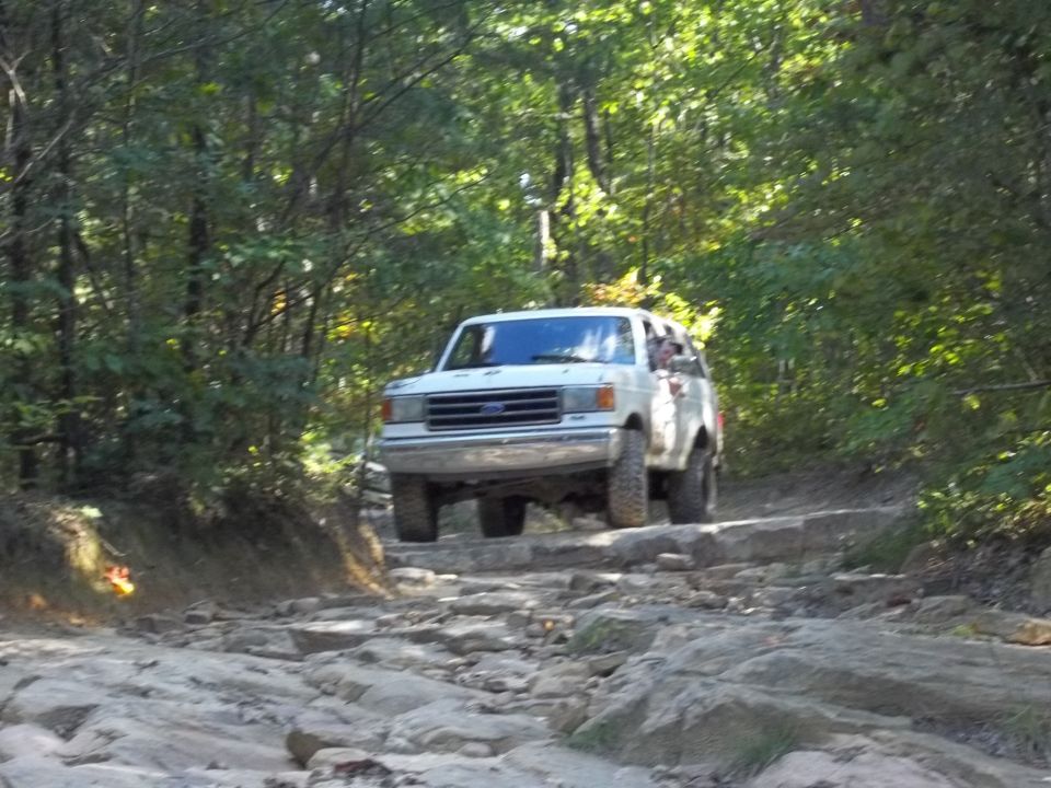 White Ford Bronco driving over rocks on a wooded trail. The vehicle appears to be a 1990 model