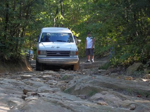 1990 Ford Bronco - A white 1990 Ford Bronco navigates a challenging rocky path in a wooded area.
