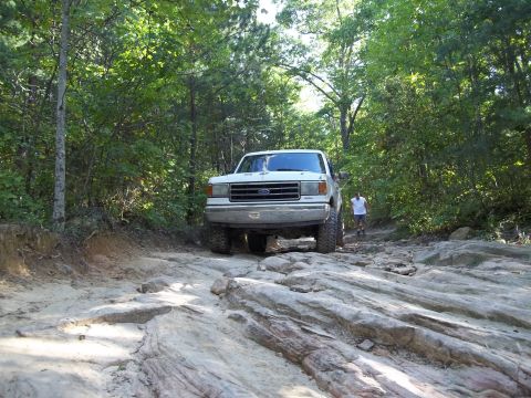 1990 Ford Bronco - A white 1990 Ford Bronco navigates a challenging rocky path through a wooded area.