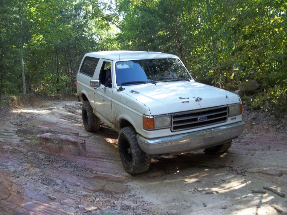1990 Ford Bronco driving on a dirt and rock trail with trees in the background