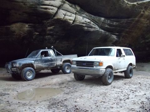 1990 Ford Bronco - Two off-road vehicles, a white Ford Bronco and a black Toyota pickup, are parked side-by-side in a natural rock formation.