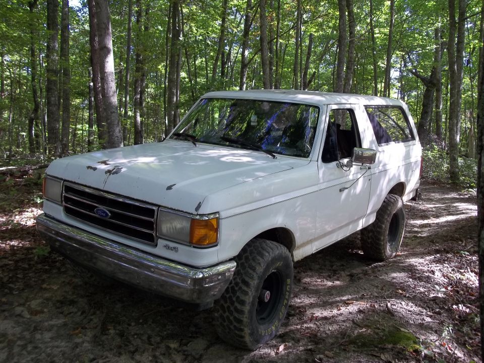 White 1990 Ford Bronco parked on a dirt path in a forest