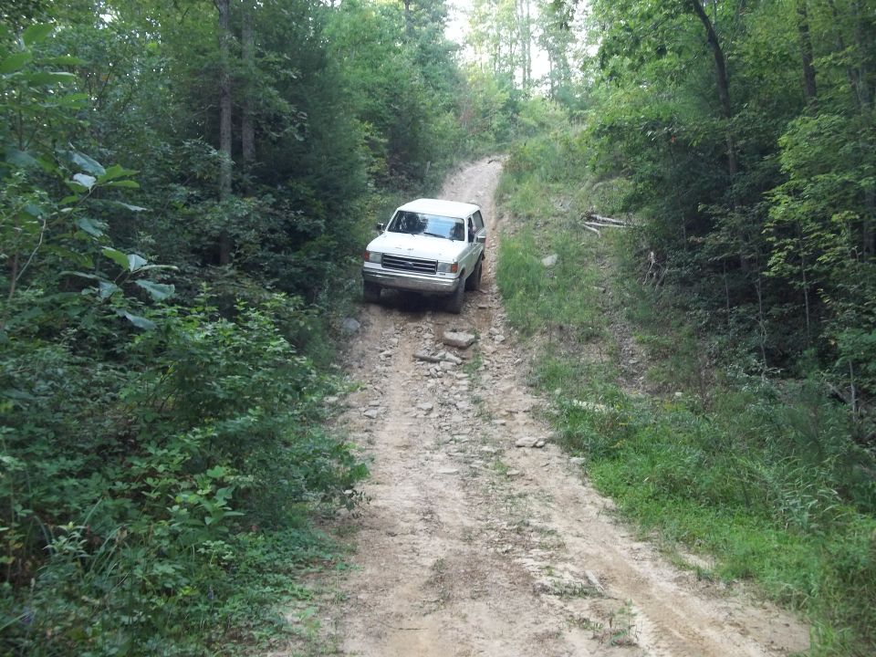 White 1990 Ford Bronco driving on a rocky dirt road through woods