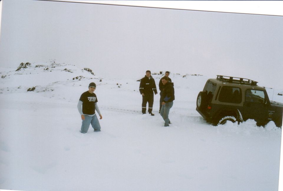 Dark 4x4 SUV with roof rack stuck in deep snow, four people standing nearby