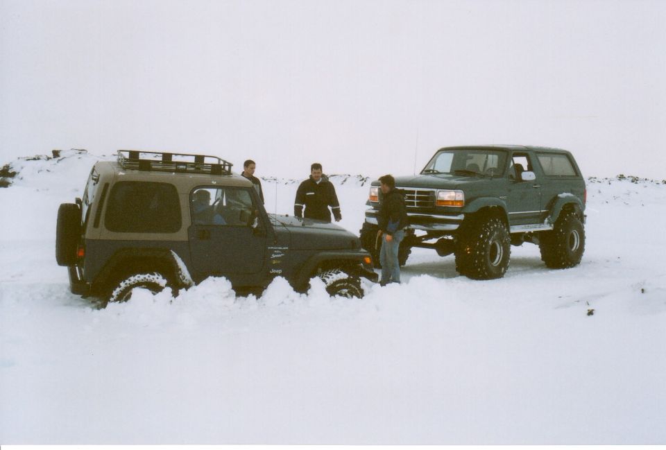 Lifted dark Bronco and tan-top Jeep Wrangler parked in deep snow with three men standing between them