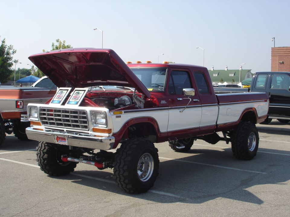 Lifted red and white pickup truck with hood open, showing engine. Two framed pictures sit on top of the engine