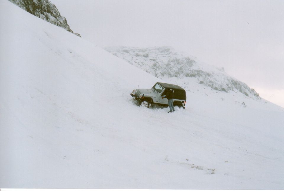 Light SUV with dark top stuck in deep snow on a steep slope, person standing beside it