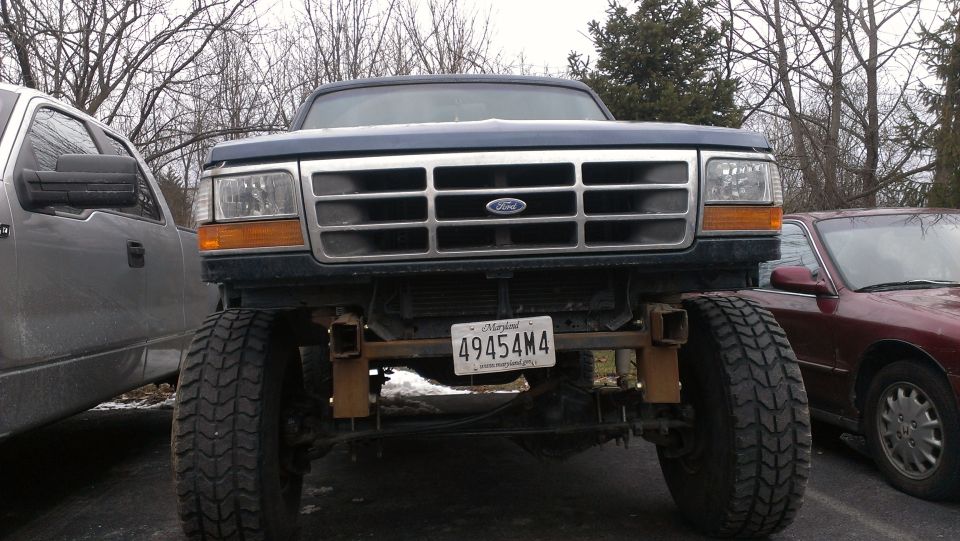 Front view of a lifted dark blue Ford truck with a chrome grille and a Maryland license plate. Another truck is to the left and a sedan to the right