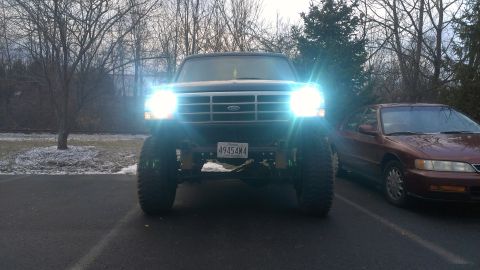 1995 Ford Bronco - A lifted black Ford Bronco is parked in front of a red sedan, its headlights shining brightly.