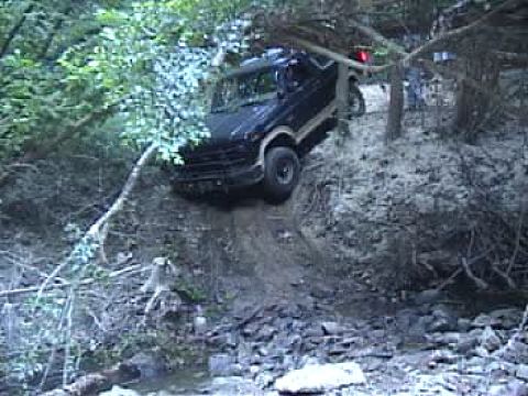 1983 Ford Bronco - A dark SUV navigates a challenging off-road trail through a wooded area.