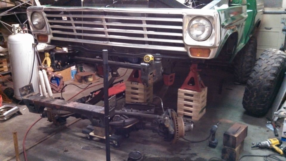 Front end of a green Ford truck on jack stands, with the grille and front axle visible. It's in a garage with tools around