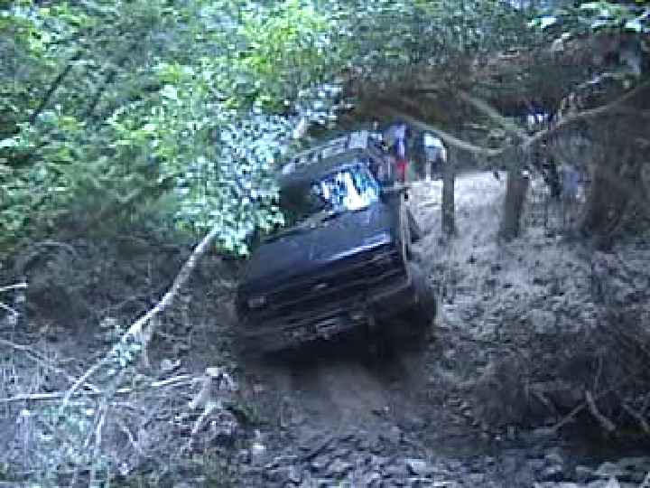 Dark SUV tilted on steep rocky forest trail with onlookers in background