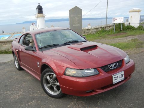 2003 Mercury Marauder - A red Ford Mustang is parked by the coast with a lighthouse and a monument in the background.