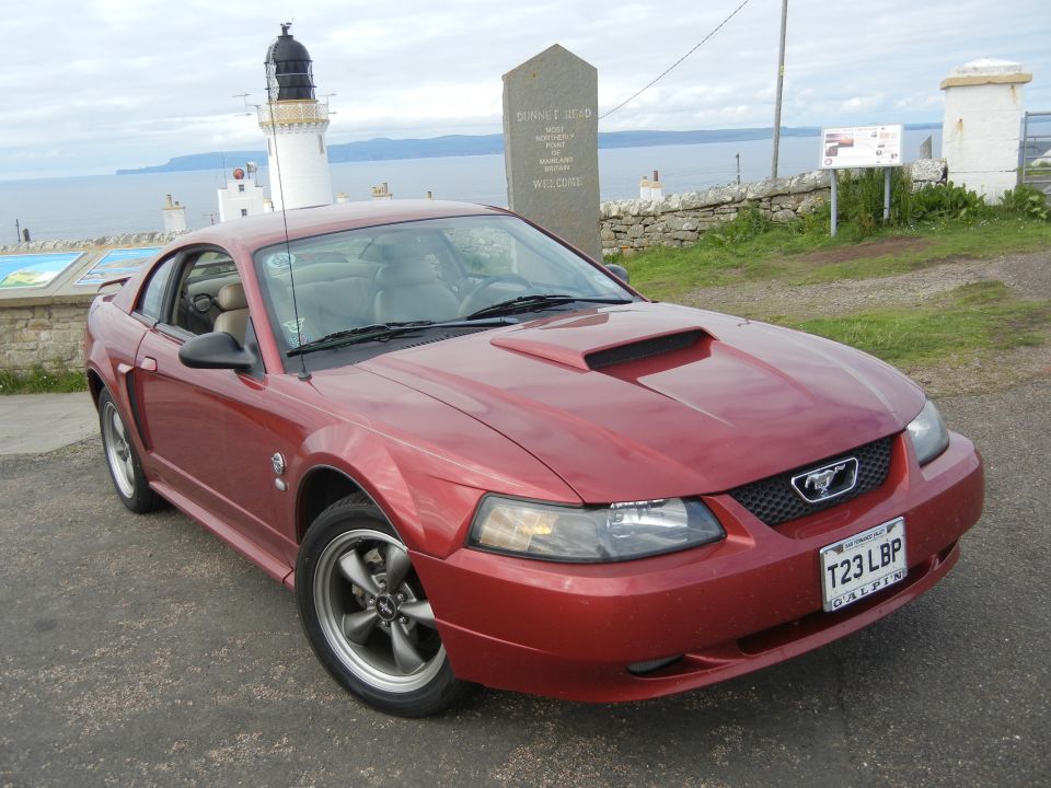 Red Ford Mustang with hood scoop parked near a lighthouse and a stone monument