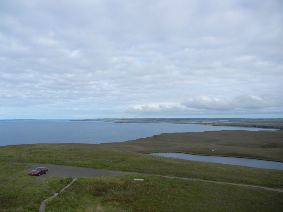 Red sedan parked on asphalt overlooking a large body of water and rolling hills