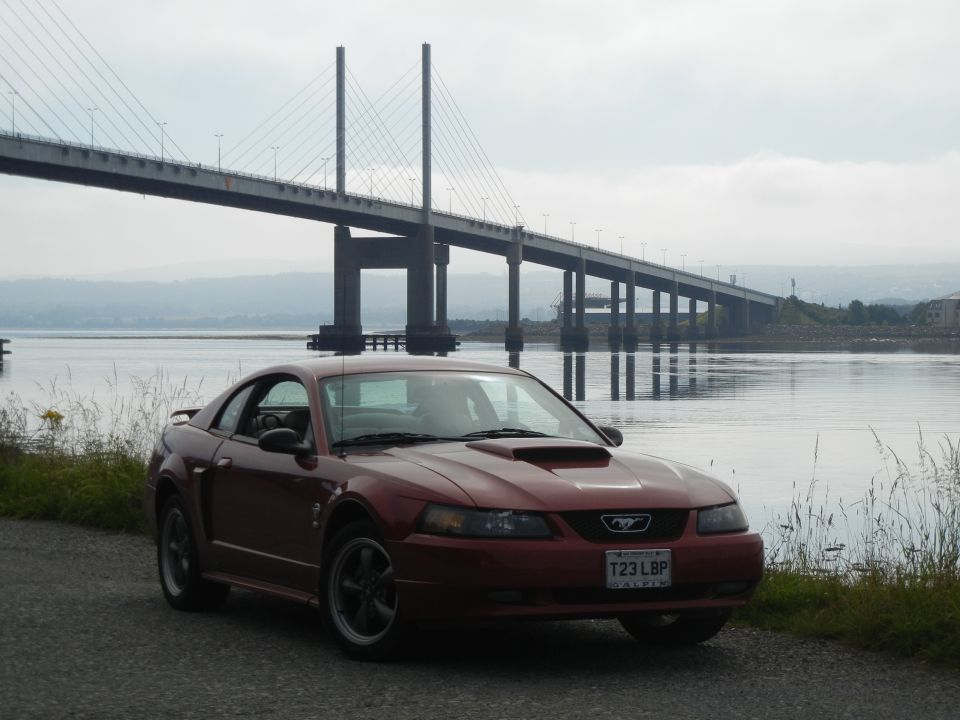 Red Ford Mustang coupe with hood scoop parked on gravel, large bridge and water in background