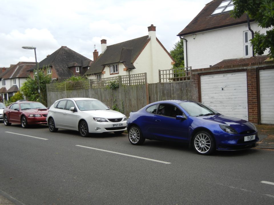Blue Ford Puma coupe parked on street next to a white Subaru Impreza and a red Ford Mustang