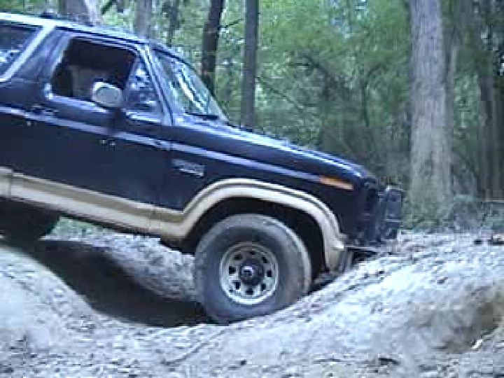 Dark blue 80s Ford Bronco with tan trim flexing into a muddy rut in the woods