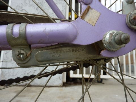 1900 Yugo GVX - Close-up of a bicycle's rear derailleur, marked 'Shimano CB-E110 Japan', attached to a purple frame.