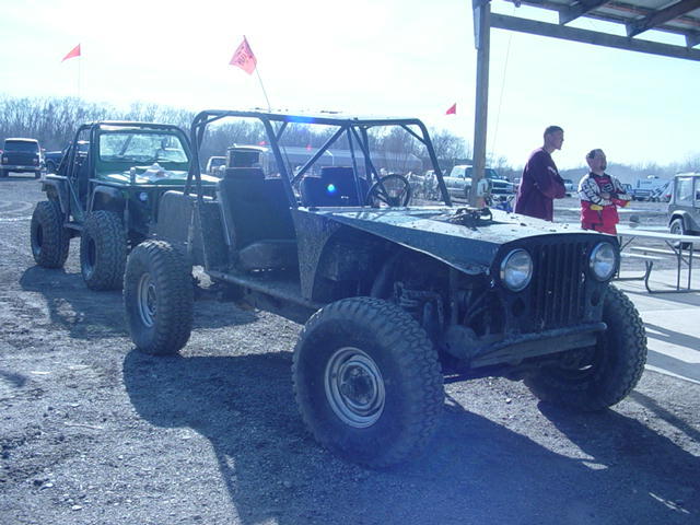 Custom off-road buggy with large tires and roll cage, parked next to another off-road vehicle. Two people stand nearby