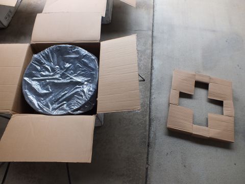 2003 Ford Crown Victoria - round plastic-wrapped alloy wheel sits inside an open box; a cut cardboard insert lies on the floor beside it.