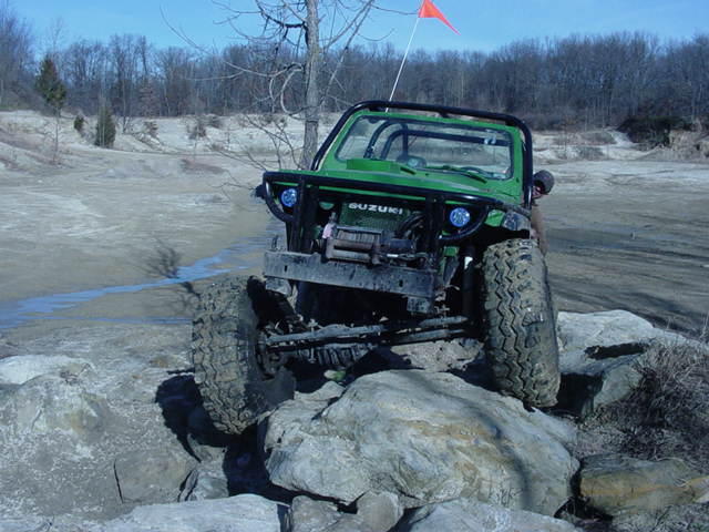 Green Suzuki Samurai with off-road tires climbing over rocks, orange flag on antenna