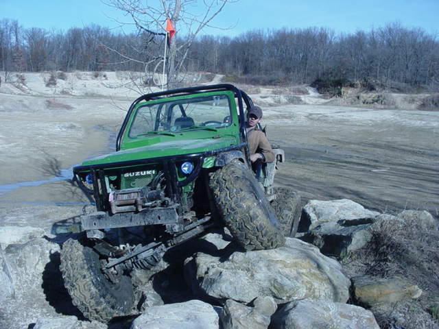 Green Suzuki Samurai with a roll cage is driving over rocks. Driver is visible