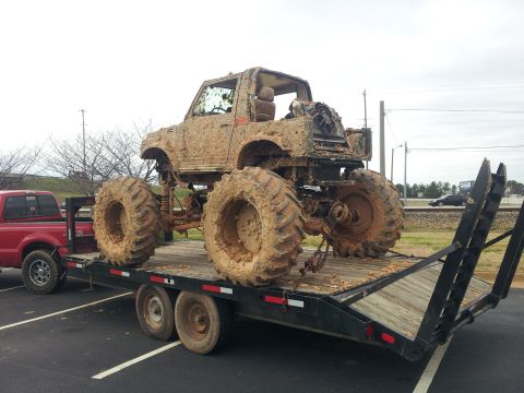 1986 Ford Bronco - Mud-covered off-road vehicle on a trailer, towed by a red pickup truck.