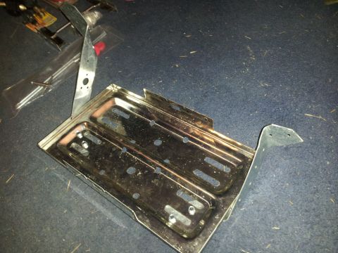1986 Ford Bronco - A metal battery tray with mounting brackets is shown on a dark carpeted surface.