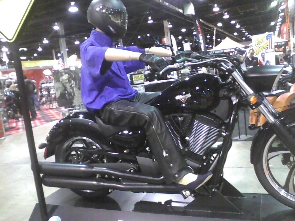 Black motorcycle with a mannequin wearing a purple shirt and black leather pants, sitting on it. The mannequin has a black helmet