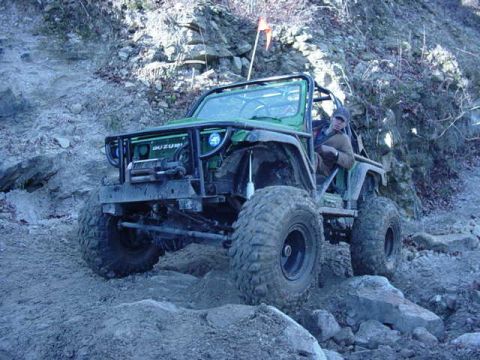 1992 Ford Bronco - A green off-road vehicle navigates a challenging rocky incline, with a driver visible behind the wheel.