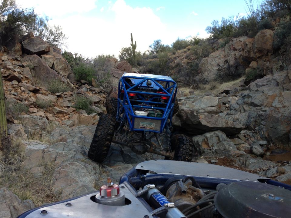 Blue off-road buggy climbing rocky terrain, another vehicle's hood visible in foreground