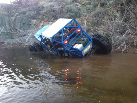 1966 Ford Bronco - Off-road buggy with blue roll cage is stuck in shallow water near a rocky bank.