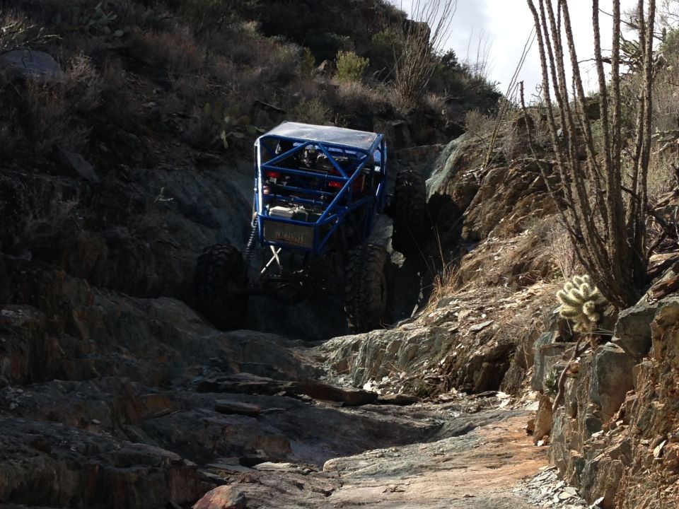 Blue off-road buggy with roll cage climbing a steep, rocky desert trail. Supermotors.net logo visible