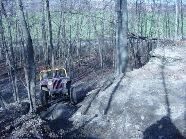 Jeep CJ-7 with yellow roll cage driving up a rocky, wooded incline