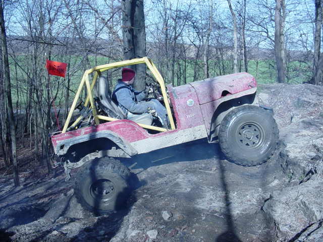 Red Jeep Wrangler with yellow roll cage climbing a rocky incline, driver wearing a blue jacket and red hat
