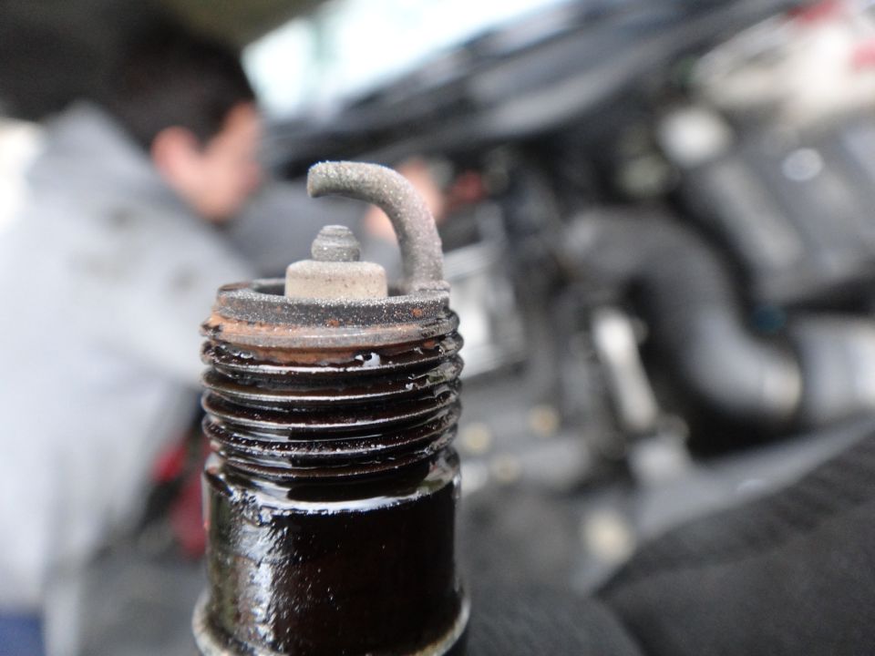 Close-up of a dirty, used spark plug with black deposits and a bent electrode, with an engine and person blurred in the background