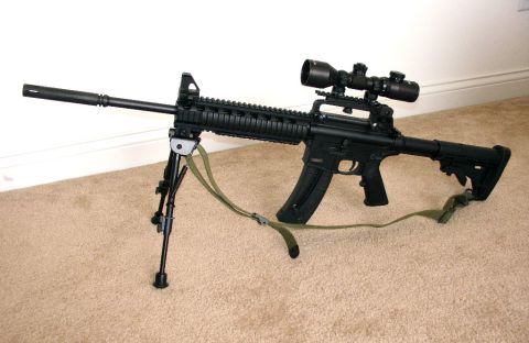 2003 Ford Crown Victoria - A black rifle equipped with a bipod and scope is displayed on a carpeted floor.