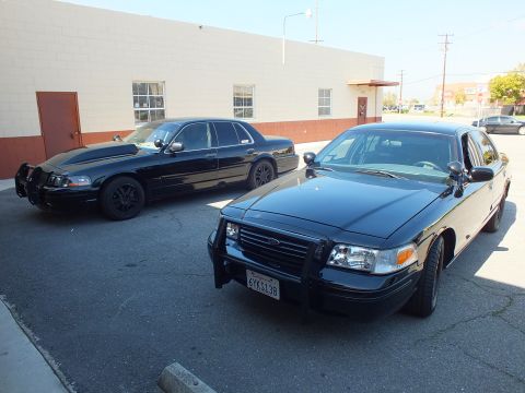 2003 Ford Crown Victoria - Two black Ford Crown Victoria Interceptor sedans with front push bars angled in a lot near a wall