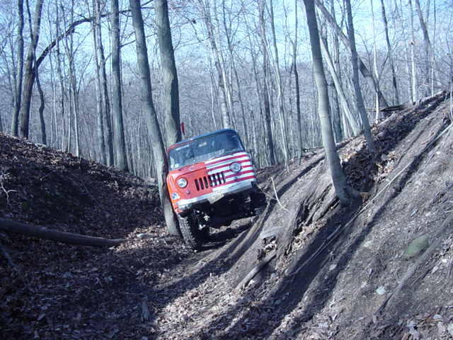Red Jeep CJ-5 climbing a steep, wooded hill. Grille indicates 1970s model