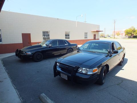 2003 Ford Crown Victoria - Pair of black Ford Crown Victoria sedans, one front-on with push bar, sit in a parking lot by a warehouse.