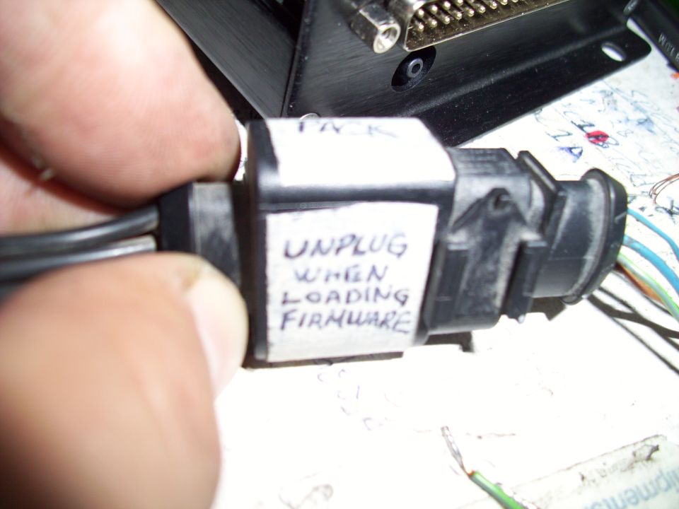 Black electrical connector with white tape labels reading 'PACK' and 'UNPLUG WHEN LOADING FIRMWARE'