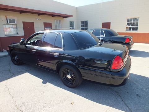 2003 Ford Crown Victoria - Sun glints off a black Crown Victoria rear quarter; a matching sedan sits nearby by the warehouse.