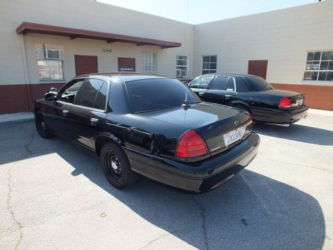 2003 Ford Crown Victoria - Back view of two black Crown Victoria sedans with steel wheels in a lot beside an industrial unit.