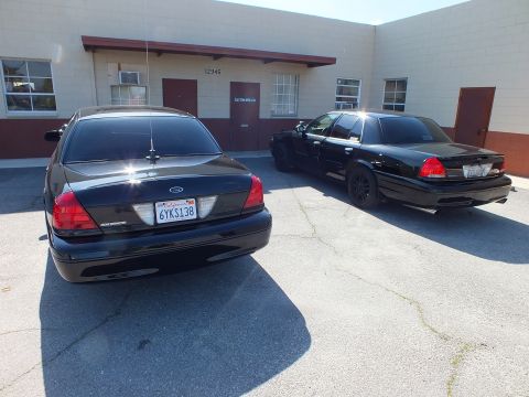 2003 Ford Crown Victoria - pair of black Crown Victoria sedans sit nose-to-tail by a warehouse, one tagged 6YKS138.
