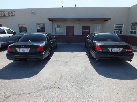 2003 Ford Crown Victoria - Pair of black Crown Victoria sedans, plates 6YKS138 and EVIL VIC, parked outside a warehouse.
