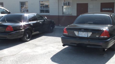 2003 Ford Crown Victoria - Close rear view of two black Crown Victoria sedans, the right one tagged EVIL VIC, at a warehouse.