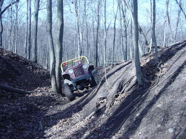 Maroon Jeep CJ-7 with yellow roll cage climbing a steep, wooded hill