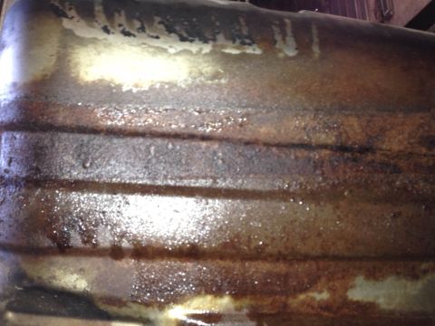 1979 Ford Bronco - Close-up of a heavily rusted metal part, possibly a fuel tank or oil pan, with visible horizontal ridges and some liquid residue.