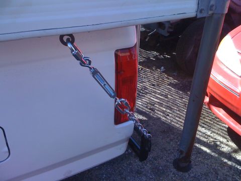 1986 Yugo GV - Chain and turnbuckle assembly secures a load to the bed of a white pickup truck, with a red vehicle visible to the right.